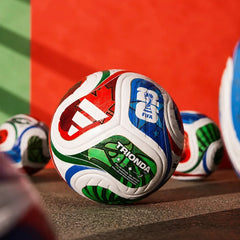 A close-up of a white soccer ball with red, green, and blue graphics, featuring the Adidas logo, FIFA World Cup branding, and the word TRIONDA, resting on a textured surface with other soccer balls blurred in the background against a red and green backdrop.