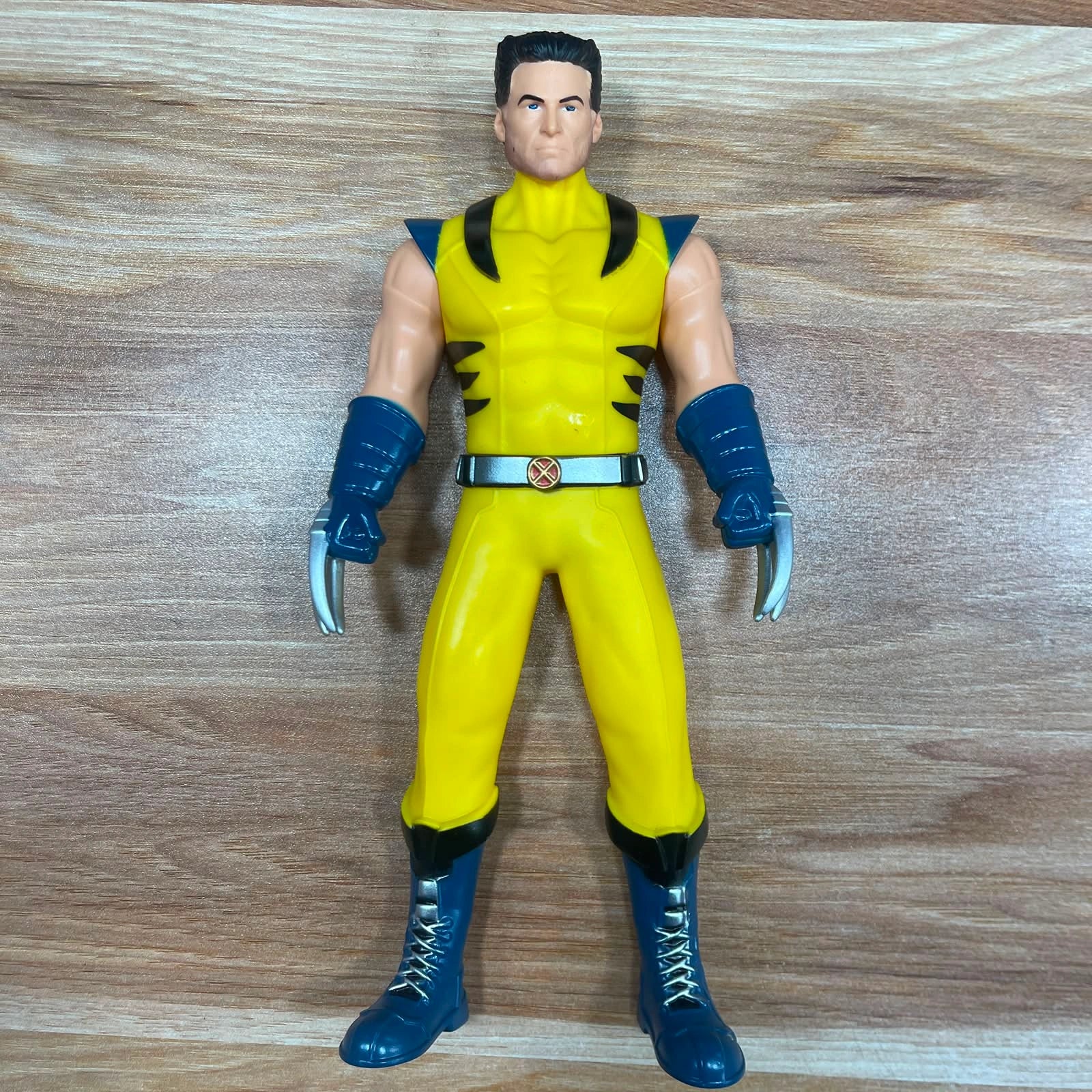 A Wolverine action figure standing on a light brown wood-grain surface. The figure is wearing his classic yellow and blue costume with black accents, silver claws, and a silver belt with a gold emblem.