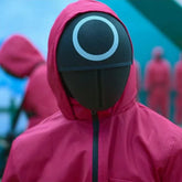 Person wearing a pink hooded jacket and a Squid Game Circle Guard Mask on a blue background.
