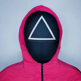 Person wearing a pink hooded jacket with a black squid game face mask featuring a white triangle symbol on a gray background