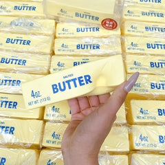 Hand holding a 4oz butter stick with stacked butter packages in the background