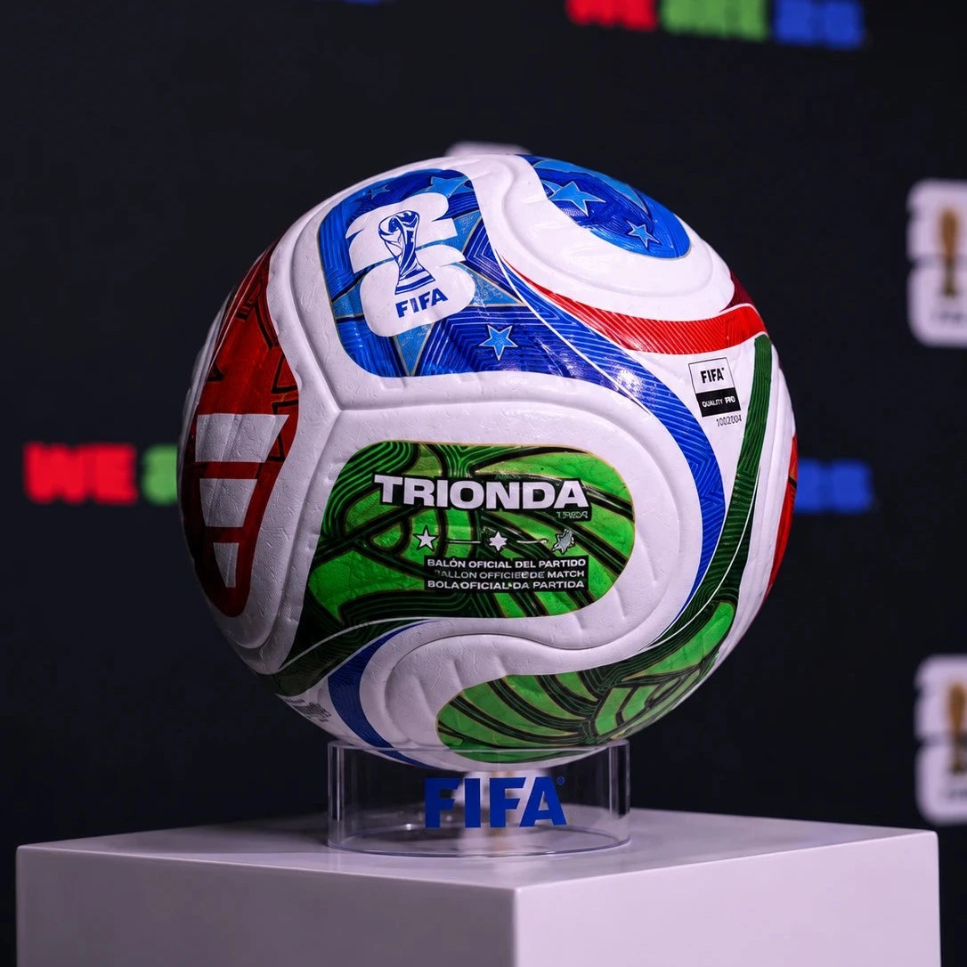 A FIFA-branded soccer ball with the word TRIONDA on the front, displayed on a clear stand atop a white pedestal against a dark backdrop with colorful blurred logos.