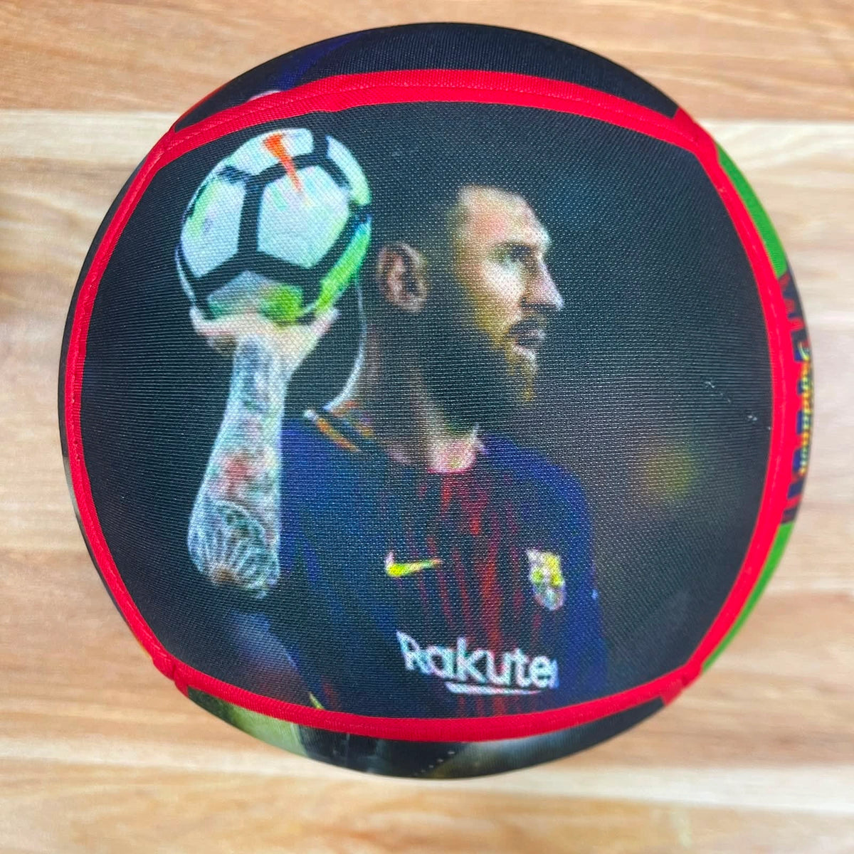 A round black cushion featuring a printed graphic of Lionel Messi in an FC Barcelona jersey holding a soccer ball, with a red border and green accents, placed on a light wood floor.