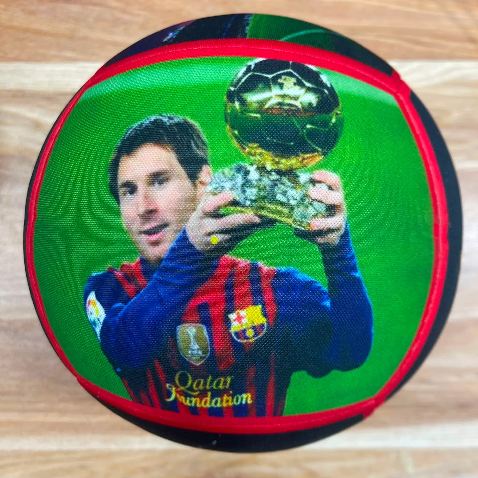 A round soccer ball featuring a printed graphic of Lionel Messi holding the FIFA World Cup trophy, with a green background, red trim, and FC Barcelona branding.