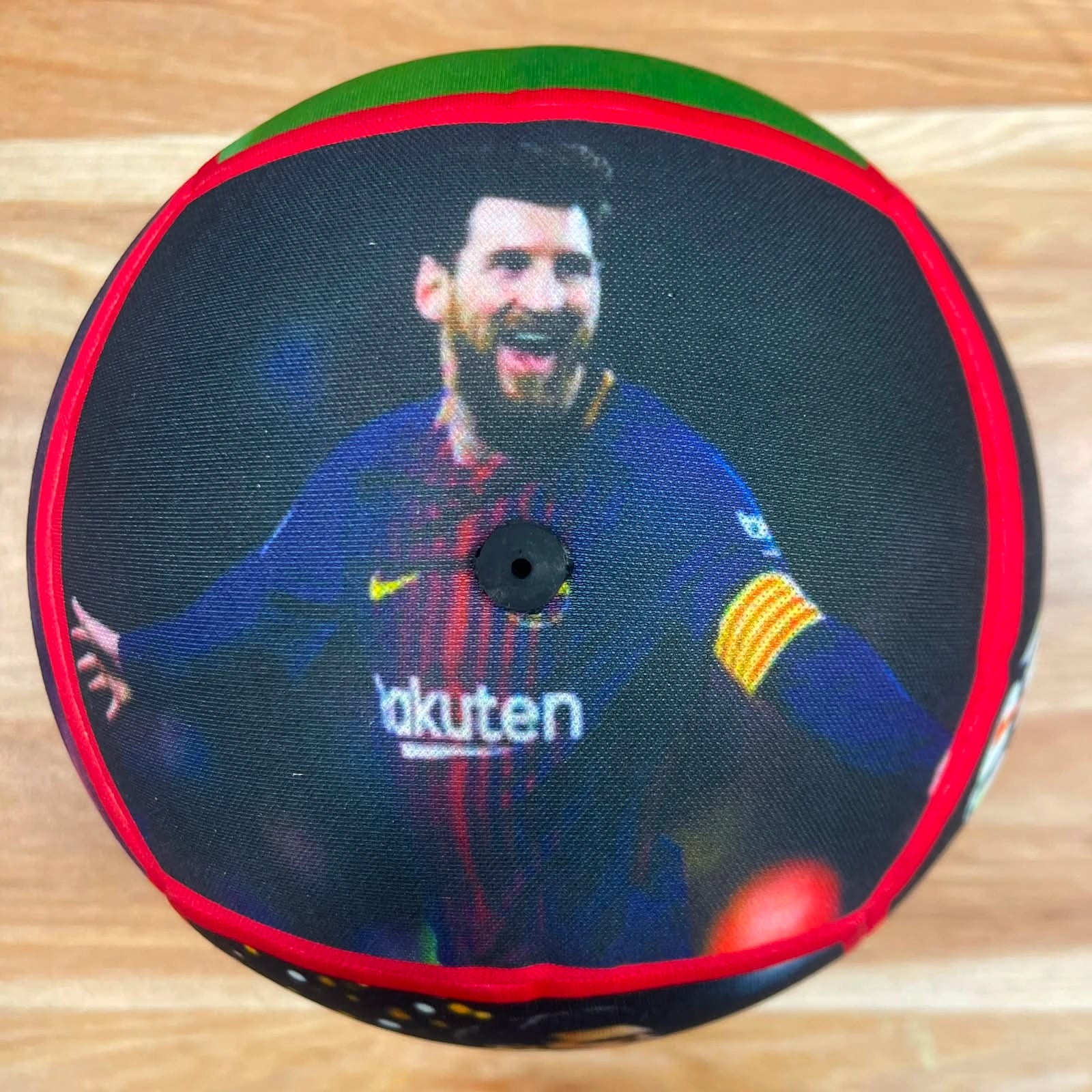 A round decorative pillow featuring a printed graphic of soccer player Lionel Messi in a blue FC Barcelona jersey, with a red and green border, photographed on a light wood floor.