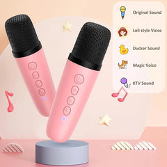 Two pink wireless microphones with black mesh tops displayed on a light blue-gray stand against a soft peach background. A white panel on the right lists voice modes including Original Sound, Loli style Voice, Ducker Sound, Magic Voice, and KTV Sound, with small decorative icons and music note graphics around the products.