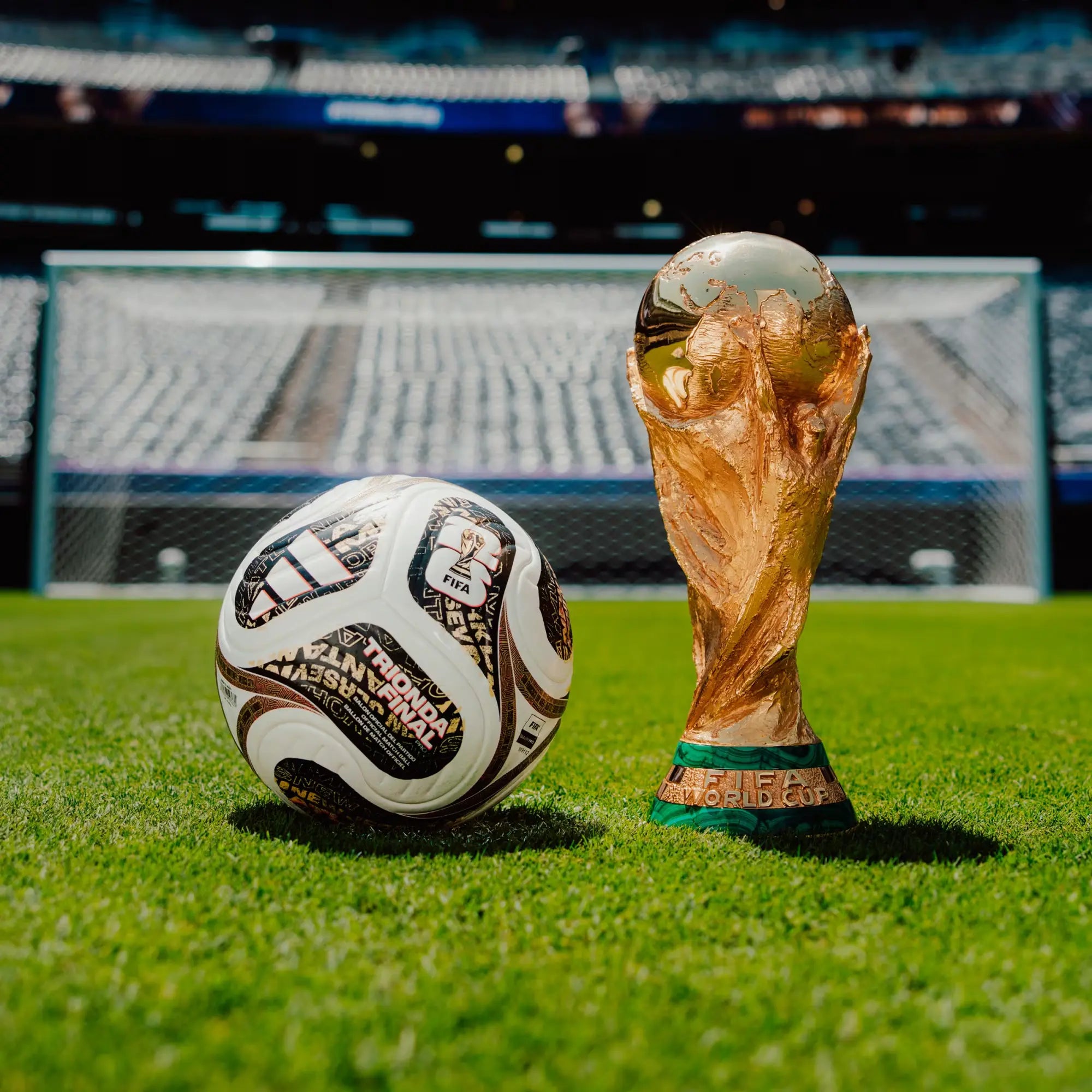 A white Adidas soccer ball and the FIFA World Cup trophy sit on a green grass field in front of a stadium with blurred seating and a goal in the background.